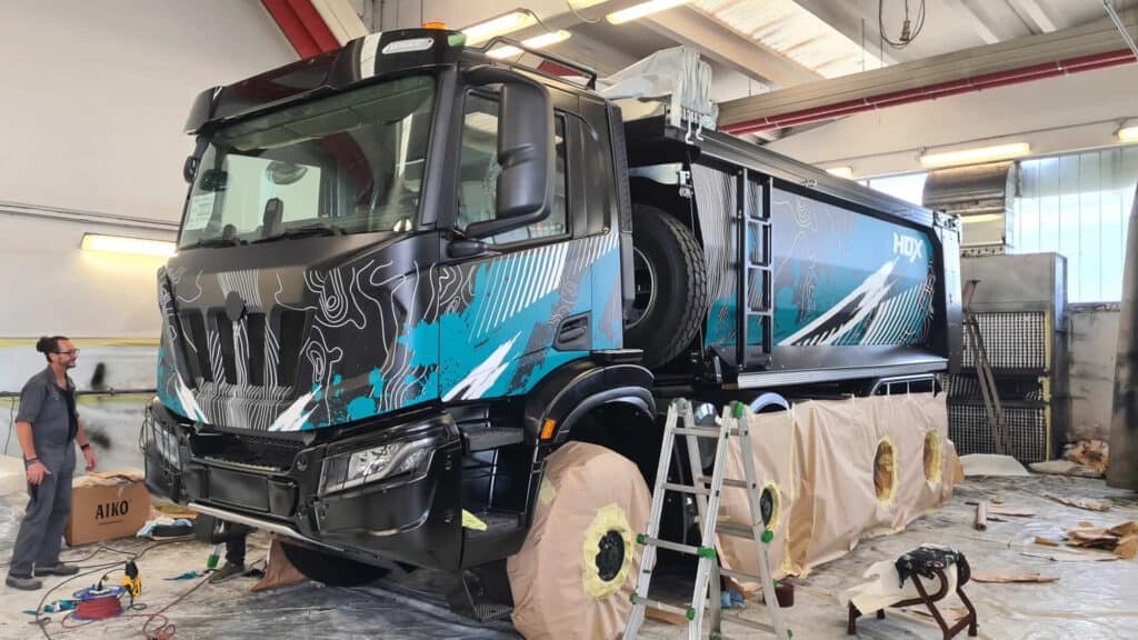 Un grande camion in un'officina sta ultimando la sua verniciatura personalizzata. Le ruote sono coperte e vari utensili e materiali sono sparsi in giro. Si vede una persona che lavora vicino al camion.