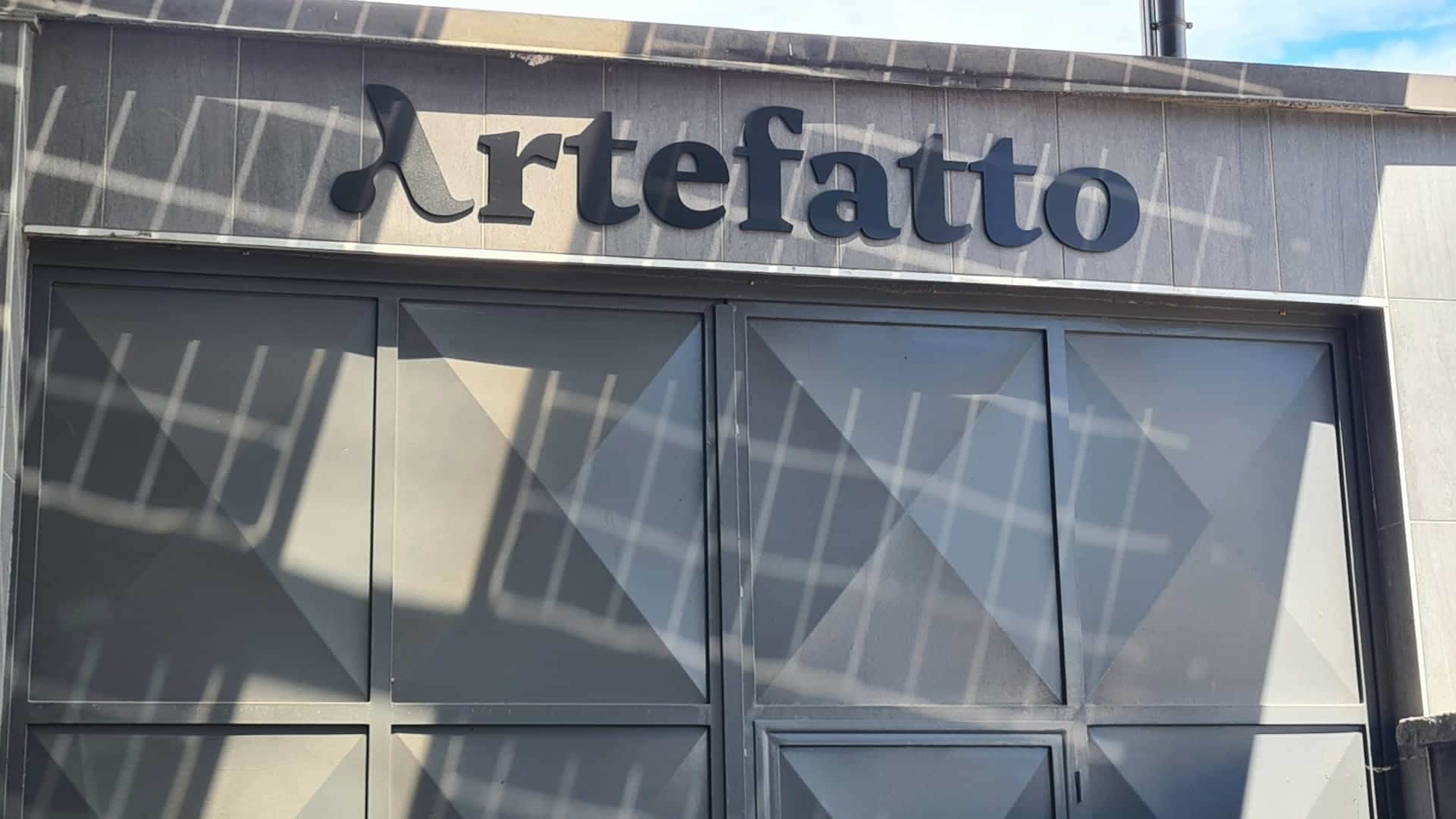 Il logo "Artefatto" esposto in lettere in grassetto e sorprendenti sopra imponenti porte grigie, esaltate da un intrigante gioco di motivi d'ombra. In Modoprint, siamo specializzati nel rendere la tua segnaletica esterna non solo visibile, ma indimenticabile.